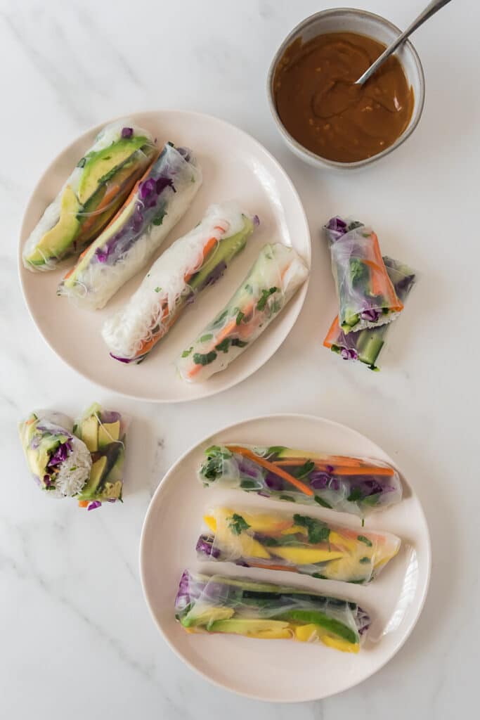 overhead view of two white plates with three spring rolls on each plate, with two spring rolls that have been cut in half and stacked on top of each other on the side, and bowl of peanut dippgin sauce