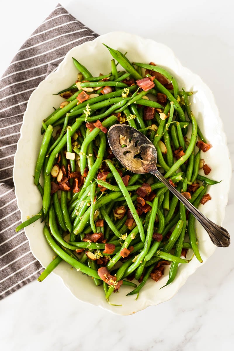 overhead view of green bean almondine in white platter with serving spoon and napkin beside