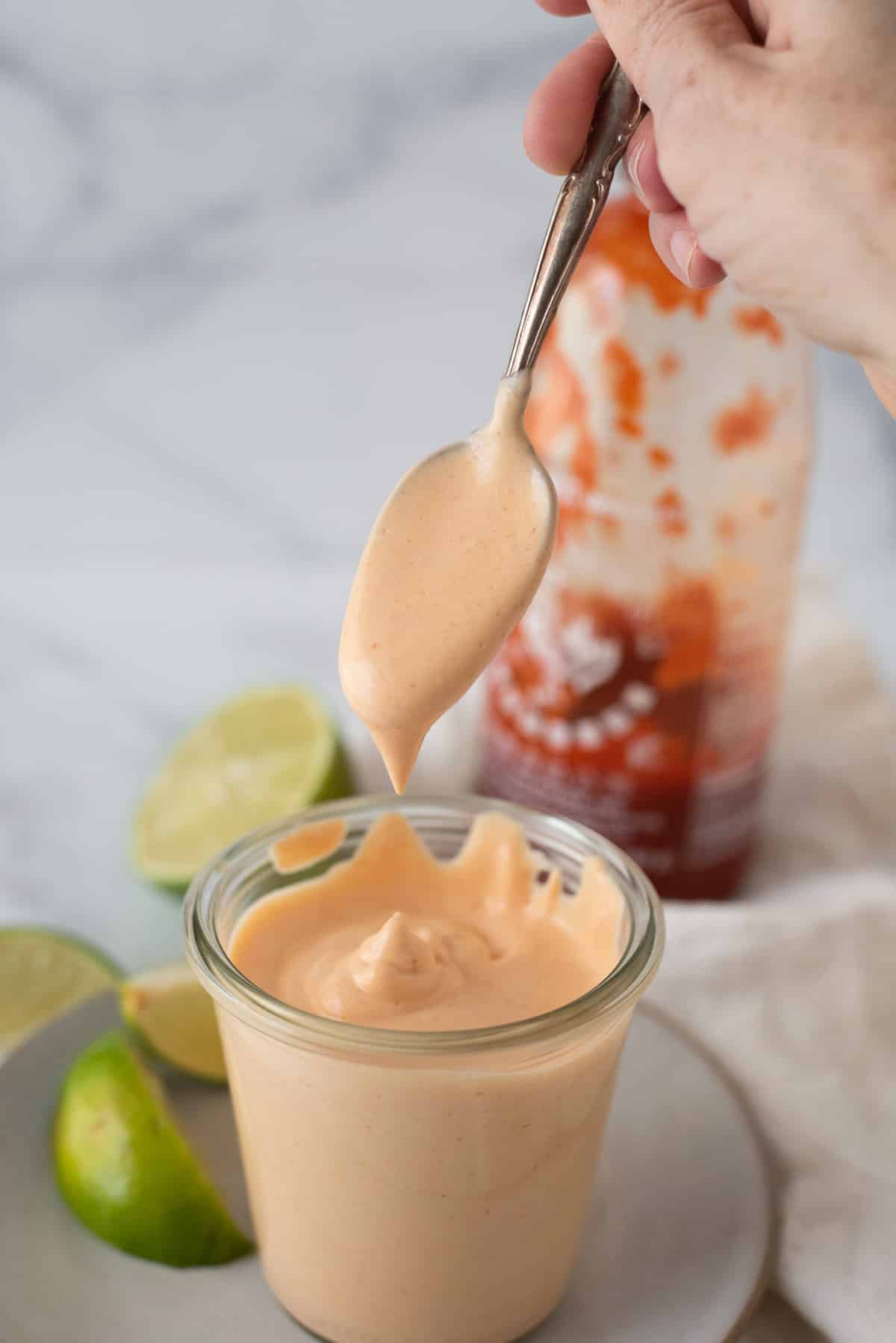 glass container filled with spicy mayo, with spoon taking some out and limes behind